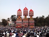 A large crowd gathers around three towering, ornate chariots adorned with intricate decorations in bright colors. The chariots are set against a backdrop of tall green trees under a clear sky.