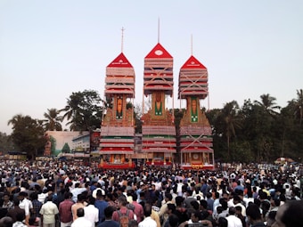 A large crowd gathers around three towering, ornate chariots adorned with intricate decorations in bright colors. The chariots are set against a backdrop of tall green trees under a clear sky.