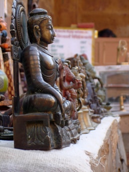 A row of intricately designed brass statues, with a prominent large statue of a seated Buddha in focus. The background is slightly blurred, showcasing more statues and a cluttered market setting.