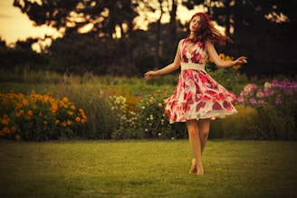 A cheerful woman in a flowing floral dress posing gracefully in a sunlit garden.