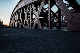 A low-angle view of a steel bridge with intricate cross-bracing and riveted beams. The texture and details of the metalwork are highlighted against a backdrop of a dim evening sky.