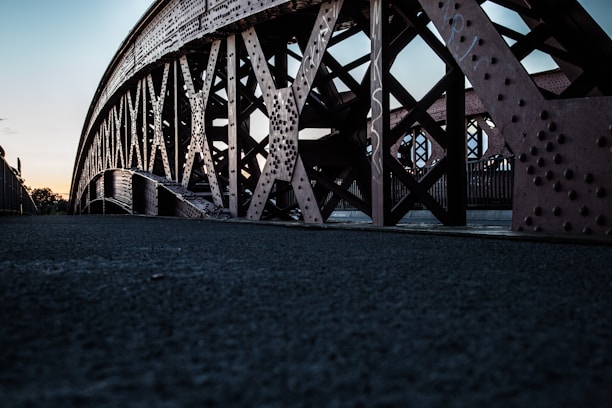 A low-angle view of a steel bridge with intricate cross-bracing and riveted beams. The texture and details of the metalwork are highlighted against a backdrop of a dim evening sky.