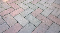 A close-up view of an interlocking brick pavement with a herringbone pattern. The bricks are a mix of gray and red colors, and there are small patches of green moss or grass growing between some of the bricks.