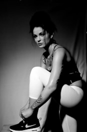 A woman is dressed in athletic wear, including a tank top, white leggings, and black Nike sneakers. She is kneeling on one knee, with the other leg bent and foot on the ground. She appears to be tying her shoe. The lighting is dramatic, creating strong contrasts and shadows, with a monochrome filter.