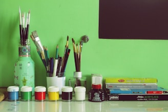A collection of painting and drawing supplies arranged neatly against a green background. There are several paintbrushes and pencils organized in containers. In front, small jars of paint in various colors such as blue, white, green, red, yellow, and black are lined up. To the right, there are boxes of watercolors and acrylics stacked. A black rectangle, possibly a blackboard or part of the wall, is visible in the upper right corner.