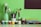 A collection of painting and drawing supplies arranged neatly against a green background. There are several paintbrushes and pencils organized in containers. In front, small jars of paint in various colors such as blue, white, green, red, yellow, and black are lined up. To the right, there are boxes of watercolors and acrylics stacked. A black rectangle, possibly a blackboard or part of the wall, is visible in the upper right corner.