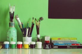 A collection of painting and drawing supplies arranged neatly against a green background. There are several paintbrushes and pencils organized in containers. In front, small jars of paint in various colors such as blue, white, green, red, yellow, and black are lined up. To the right, there are boxes of watercolors and acrylics stacked. A black rectangle, possibly a blackboard or part of the wall, is visible in the upper right corner.