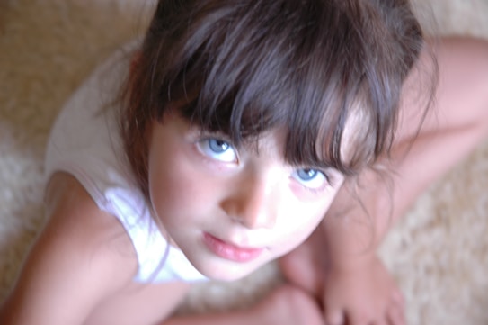 A young child with blue eyes and dark hair gazes upward, sitting with legs crossed on a light-colored, textured surface. The image is softly focused, conveying a gentle and serene atmosphere.
