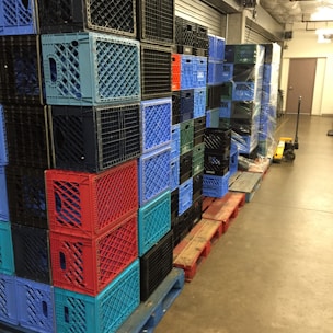 A stack of multicolored plastic crates arranged on wooden pallets in a storage area. The crates are primarily blue, red, and black, and are neatly organized in rows. A pallet jack is visible on the right side, suggesting an industrial or warehouse setting.