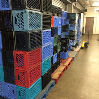 A stack of multicolored plastic crates arranged on wooden pallets in a storage area. The crates are primarily blue, red, and black, and are neatly organized in rows. A pallet jack is visible on the right side, suggesting an industrial or warehouse setting.