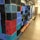 A stack of multicolored plastic crates arranged on wooden pallets in a storage area. The crates are primarily blue, red, and black, and are neatly organized in rows. A pallet jack is visible on the right side, suggesting an industrial or warehouse setting.