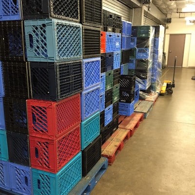 A stack of multicolored plastic crates arranged on wooden pallets in a storage area. The crates are primarily blue, red, and black, and are neatly organized in rows. A pallet jack is visible on the right side, suggesting an industrial or warehouse setting.