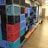 A stack of multicolored plastic crates arranged on wooden pallets in a storage area. The crates are primarily blue, red, and black, and are neatly organized in rows. A pallet jack is visible on the right side, suggesting an industrial or warehouse setting.