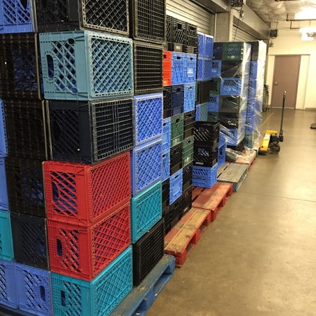 A stack of multicolored plastic crates arranged on wooden pallets in a storage area. The crates are primarily blue, red, and black, and are neatly organized in rows. A pallet jack is visible on the right side, suggesting an industrial or warehouse setting.