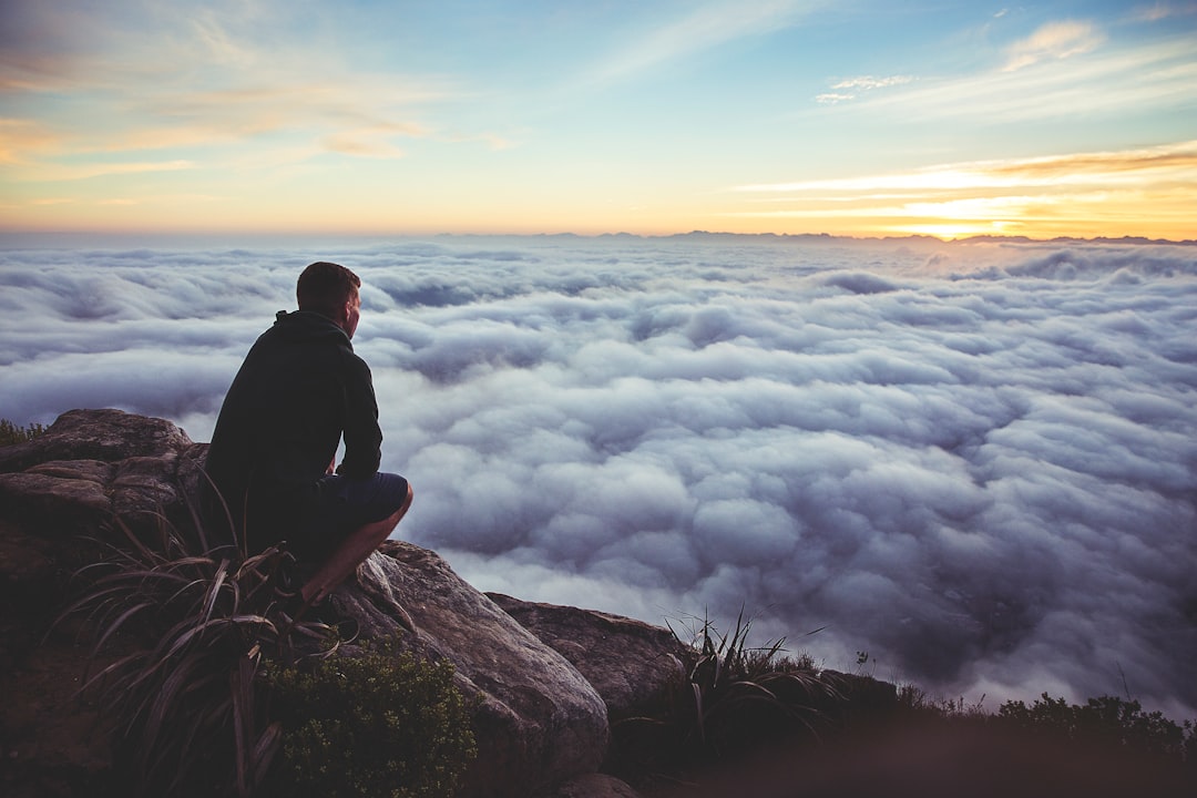 man sitting on gray rock while staring at white clouds