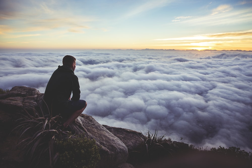 man sitting on gray rock while staring at white clouds