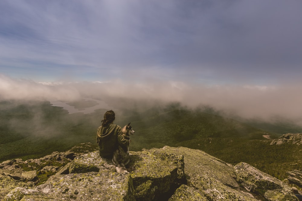 Frau und Hund sitzen auf grauer Betonfelsformation vor Berg mit Nebel