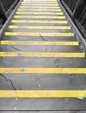 A set of wooden stairs with bright yellow anti-slip strips on each step. The stairs are flanked by wooden railings on both sides, and the surface shows some signs of weathering and natural debris like twigs and leaves.