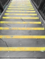 A set of wooden stairs with bright yellow anti-slip strips on each step. The stairs are flanked by wooden railings on both sides, and the surface shows some signs of weathering and natural debris like twigs and leaves.