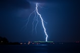 A bright, white lightning bolt striking down from a dark, cloudy sky over a body of water. The nighttime setting reveals a distant bridge illuminated by green and orange lights, reflecting slightly on the water below.