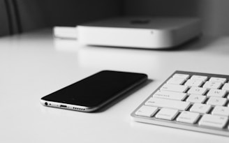A smartphone lies flat on a clean white surface next to part of a modern white keyboard. In the background, there is an electronic device, possibly a compact computer or media player, also in white.