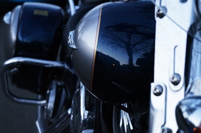 A close-up view of a motorcycle, focusing on its shiny, dark-colored fuel tank that reflects the silhouette of a tree and a building. The polished metal surfaces and parts of the engine add intricate details to the composition.