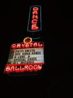 A neon sign glows brightly at night, featuring the word 'DANCE' vertically in red letters bordered by blue lighting. Below it, the neon sign reads 'CRYSTAL BALLROOM' in red, with a list of upcoming events including artists like Patti Smith and G-Love & Special Sauce. The sign is illuminated against a dark background.