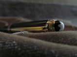 A luxury pen with a glossy black and gold finish, lying on a textured surface. The pen features intricate details, including a shiny cap with a distinctive emblem at the end.