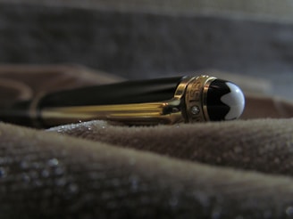 A luxury pen with a glossy black and gold finish, lying on a textured surface. The pen features intricate details, including a shiny cap with a distinctive emblem at the end.