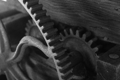 A close-up black and white image of interlocking metal gears positioned at an angle. The gears have visible texture and are set against a wooden background. Lighting highlights the detailed structure of the gear teeth.