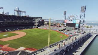 A sunlit baseball stadium with empty stands and a well-maintained green field. The outfield and large electronic scoreboard are visible, along with various advertisements. The stadium is adjacent to a body of water, with boats docked in the harbor and a bridge visible in the background. The sky is clear with a few scattered clouds.