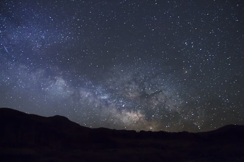 A vast, star-filled night sky with a dense cluster of stars forming the Milky Way galaxy prominently visible. The dark silhouette of a landscape stretches along the bottom edge, contrasting with the brightness of the celestial display. A faint meteor streaks across the upper left portion, adding a dynamic element to the serene scene.