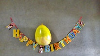 A colorful 'Happy Birthday' banner with cartoon characters is hung on a gray wall. A large yellow balloon is positioned above the word 'Birthday'. The letters are multicolored and adorned with images of a well-known cartoon character.