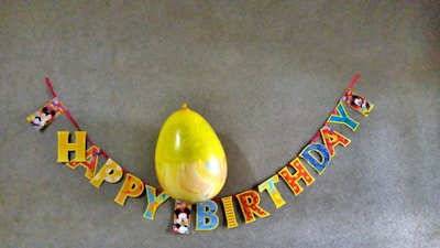 A colorful 'Happy Birthday' banner with cartoon characters is hung on a gray wall. A large yellow balloon is positioned above the word 'Birthday'. The letters are multicolored and adorned with images of a well-known cartoon character.