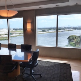 A conference room featuring a large wooden table surrounded by office chairs is seen. The room has large windows offering a panoramic view of a river and highway with multiple cars. Across the river, a city skyline is visible under a partly cloudy sky. The room is softly illuminated by wall-mounted lights.