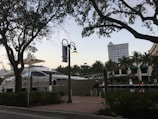 A large yacht is docked at a marina, surrounded by lush palm trees and buildings in the background. The scene is framed by silhouetted branches of nearby trees against a sky transitioning to dusk. A street lamp is mounted on a post along a brick-paved walkway that runs parallel to the water.