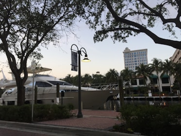A large yacht is docked at a marina, surrounded by lush palm trees and buildings in the background. The scene is framed by silhouetted branches of nearby trees against a sky transitioning to dusk. A street lamp is mounted on a post along a brick-paved walkway that runs parallel to the water.