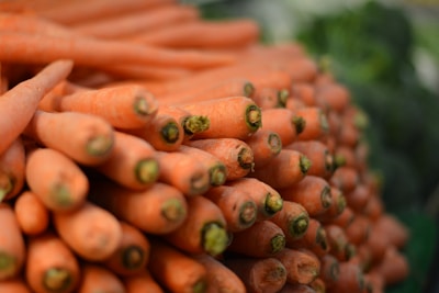 A large pile of fresh orange carrots, all aligned and stacked neatly. The focus is on the vibrant ends of the carrots, with their slightly rough textures and green stems still attached. The background is softly blurred, suggesting a market or natural setting.