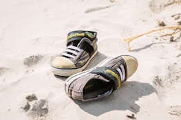 A pair of worn children's sneakers lies on a sandy surface. The sneakers are black with white laces and have a cartoon logo on the strap. The sand is fine and light-colored, with some footprints around.