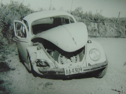 A damaged vintage car with significant frontal impact, evident by the crumpled front hood and visible tire, is stationary on a dirt path. The driver's side door is open, and the surroundings are natural with shrubbery in the background.