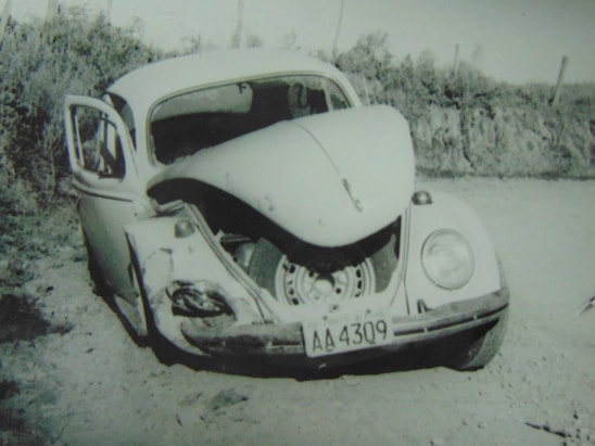 A damaged vintage car with significant frontal impact, evident by the crumpled front hood and visible tire, is stationary on a dirt path. The driver's side door is open, and the surroundings are natural with shrubbery in the background.