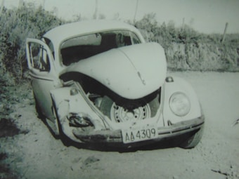 A damaged vintage car with significant frontal impact, evident by the crumpled front hood and visible tire, is stationary on a dirt path. The driver's side door is open, and the surroundings are natural with shrubbery in the background.