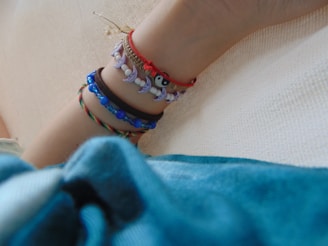 A series of colorful bracelets adorn an arm, featuring beads and charms with various designs. The background is a neutral textured surface, and a portion of a blue fabric is visible at the bottom of the frame.