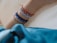 A series of colorful bracelets adorn an arm, featuring beads and charms with various designs. The background is a neutral textured surface, and a portion of a blue fabric is visible at the bottom of the frame.