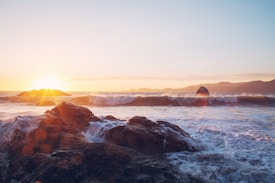 The image captures a picturesque seascape with waves crashing against rocky formations during sunset. The sun partially submerged on the horizon emits a warm golden light, illuminating the clouds and casting a soft glow over the water. Distant hills are visible along the coastline, adding depth to the scene.