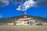 A gas station and mini mart with a cafe sign is situated in a rural area. An RV and cars are parked near the station. The backdrop includes a lush, forested hill under a bright blue sky with clouds.