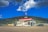 A gas station and mini mart with a cafe sign is situated in a rural area. An RV and cars are parked near the station. The backdrop includes a lush, forested hill under a bright blue sky with clouds.