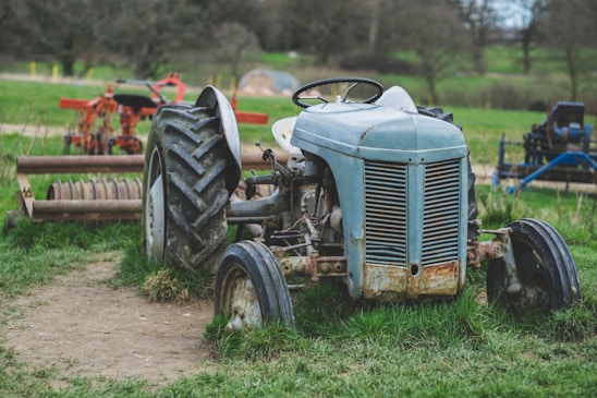 An old, weathered tractor is parked on a grassy patch in a rural setting. The tractor's body is blue with visible rust and wear, particularly on the front grille and wheels. Its large rear wheels are muddy, indicating use on farmland. In the background, there is agricultural equipment and a landscape of green fields and trees.