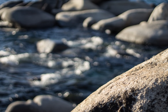 A river or stream flows over and between smooth, rounded rocks, with a focus on a closer, textured rock in the foreground. The bokeh effect creates a soft, blurred background.