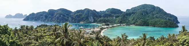 A lush, green tropical island with a vibrant blue lagoon is surrounded by rocky hills. Palm trees are abundant in the foreground, and the sea is calm with several boats visible. A small town nestles along the coastline.